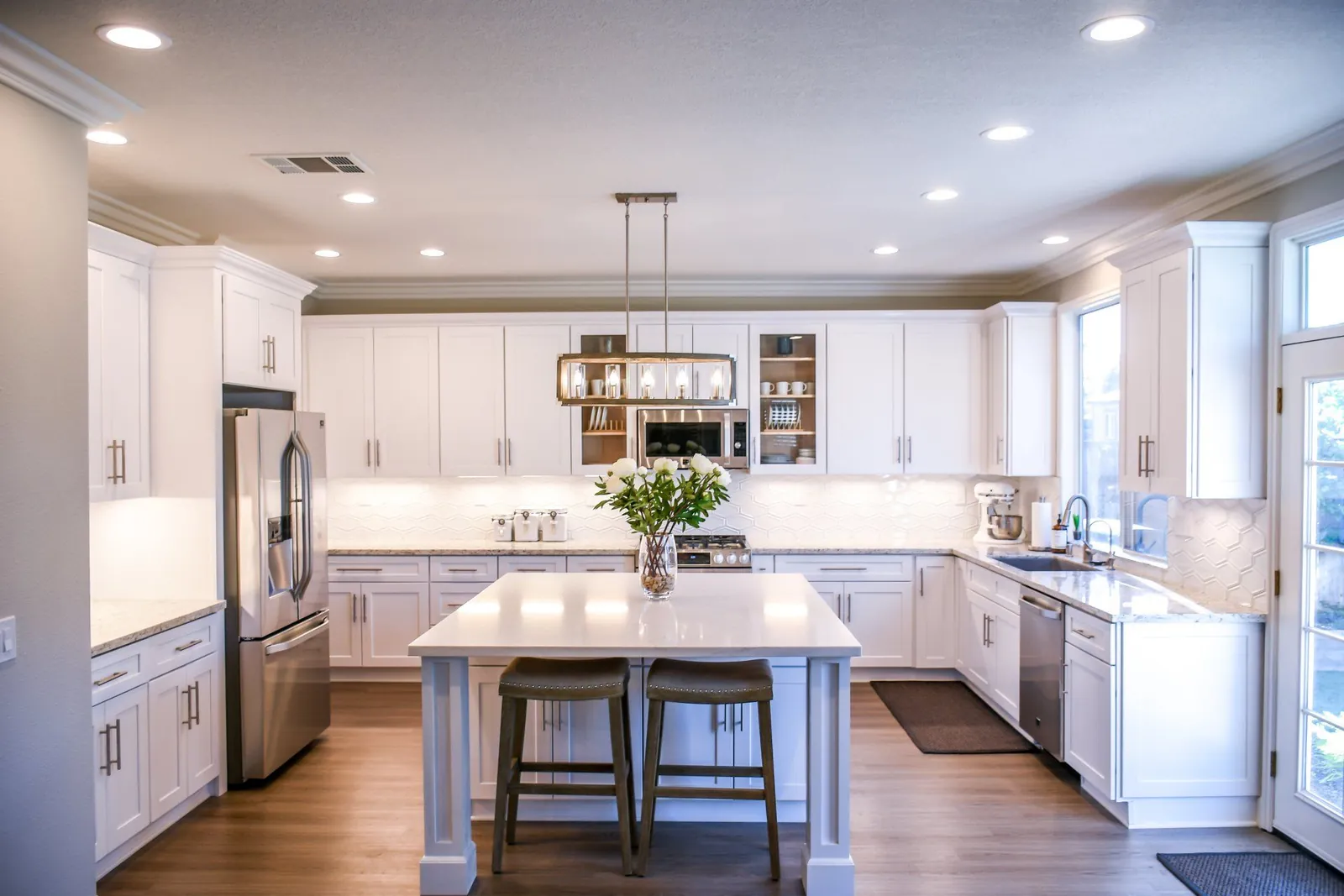 A bright, modern kitchen featuring white cabinets, a marble-topped island with two stools, and stainless steel appliances.