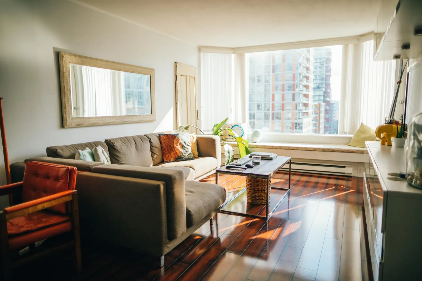 A bright living room with a large gray sectional sofa, an orange armchair, a coffee table, and a mirror on the wall.