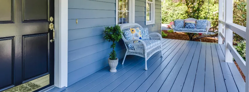 Blue front porch with a white wicker chair, potted topiary, and a porch swing, next to a black front door.