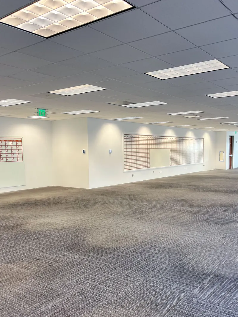 A wide, empty office space with gray carpet, white walls, drop ceiling with rectangular lights, and wall art.