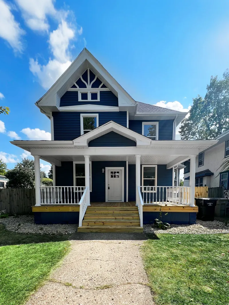 Two-story blue house with white trim, a front porch, wood steps, and a gravel walkway under a blue sky.