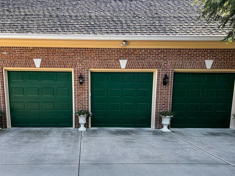 A red brick house exterior features three dark green garage doors and two white planters on the concrete driveway.