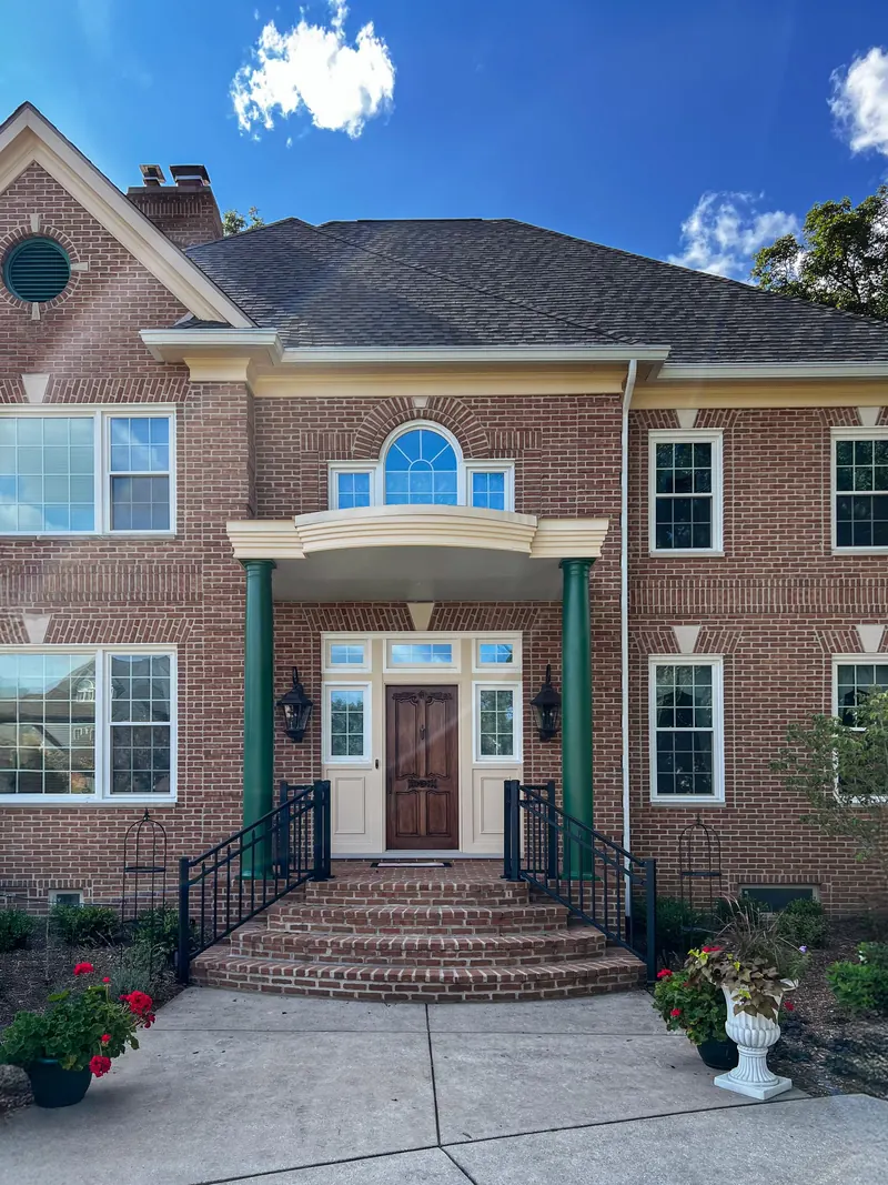 A two-story brick house with a front porch, green columns, arched window, and stairs leading to a wood front door.