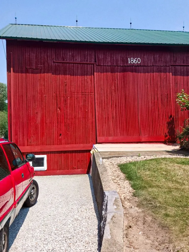A bright red barn with a green metal roof and the number 1560 on the side, next to a red car on a gravel driveway.