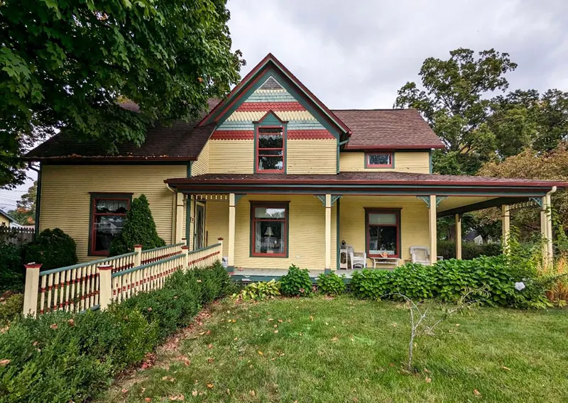 A two-story, yellow Victorian-style farmhouse with a wrap-around porch and a wheelchair ramp, set amidst green trees.