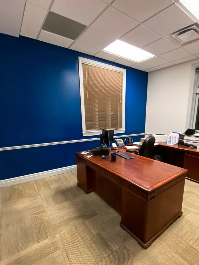 An office featuring a wooden desk against a vibrant blue wall with a window covered by blinds and light-colored carpet.