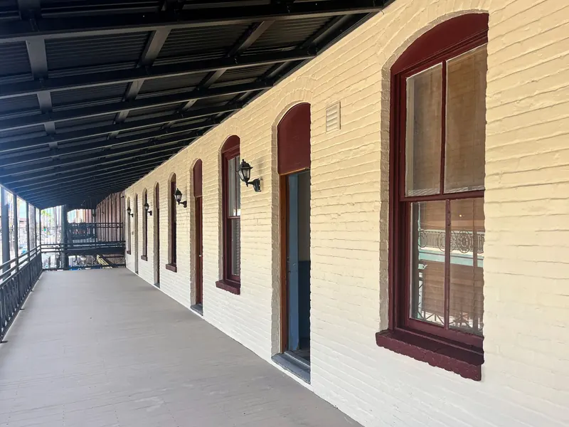 A beige brick building exterior with a long covered porch, dark red trim on windows and doors, and metal railings.