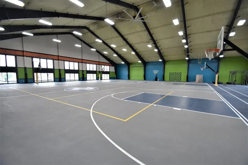 A brightly lit indoor gymnasium with gray flooring, green and blue accent walls, and basketball hoops.