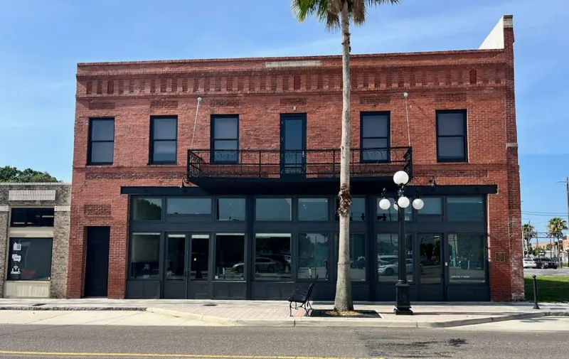 Two-story red brick commercial building with a black storefront and second-story balcony on a sunny street.