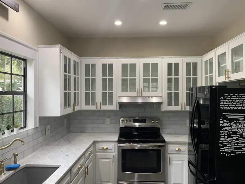 A kitchen with white cabinets featuring glass doors, light gray backsplash, marble countertops, and a black refrigerator.