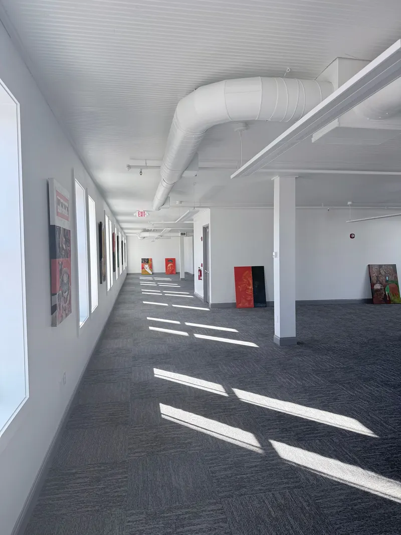 A brightly lit, modern hallway with gray patterned carpet, white walls, exposed ceiling pipes, and artwork on the walls.