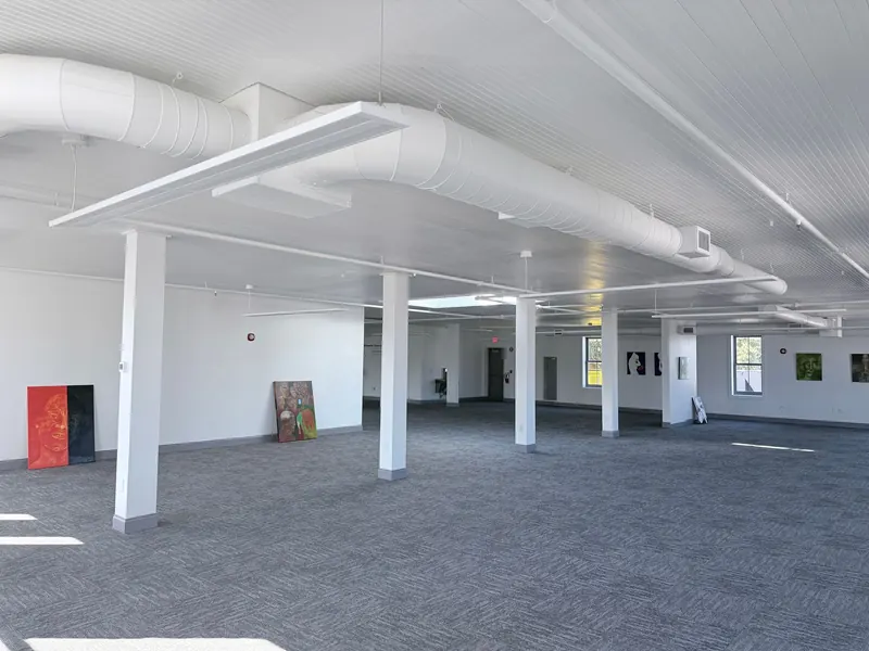 A spacious gallery interior with gray carpet, white pillars, exposed white ductwork, and artwork displayed on white walls.