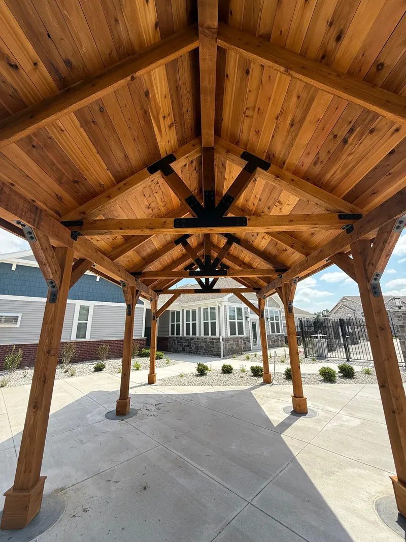 A wooden pavilion with a peaked roof and black metal structural brackets sits on a patio with a house in the background.