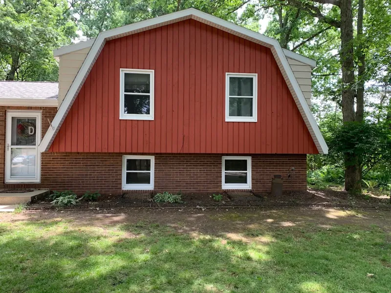 Red gambrel-roof home with white window trim and a brick foundation, surrounded by trees.