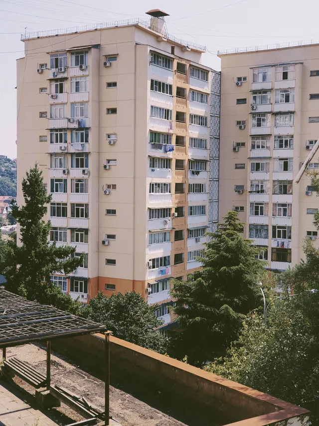 Two tall, light-tan residential apartment towers stand side-by-side behind lush green trees under a bright sky.