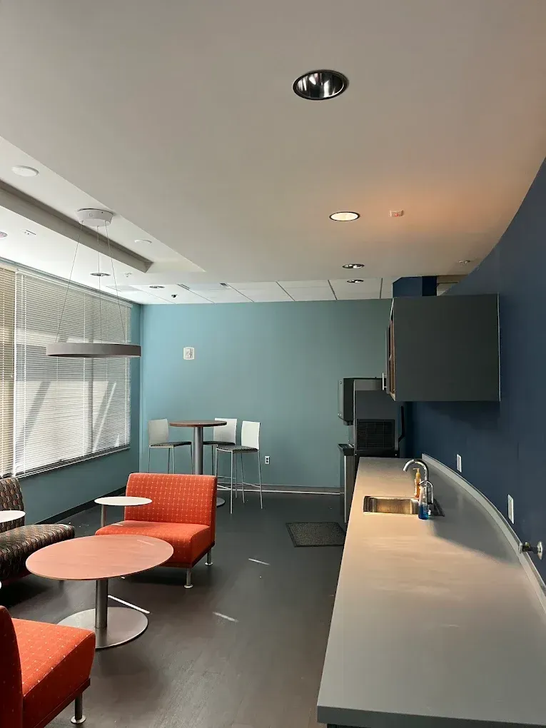 A bright office breakroom featuring orange armchairs, a round table, a high table with stools, and a long grey counter.
