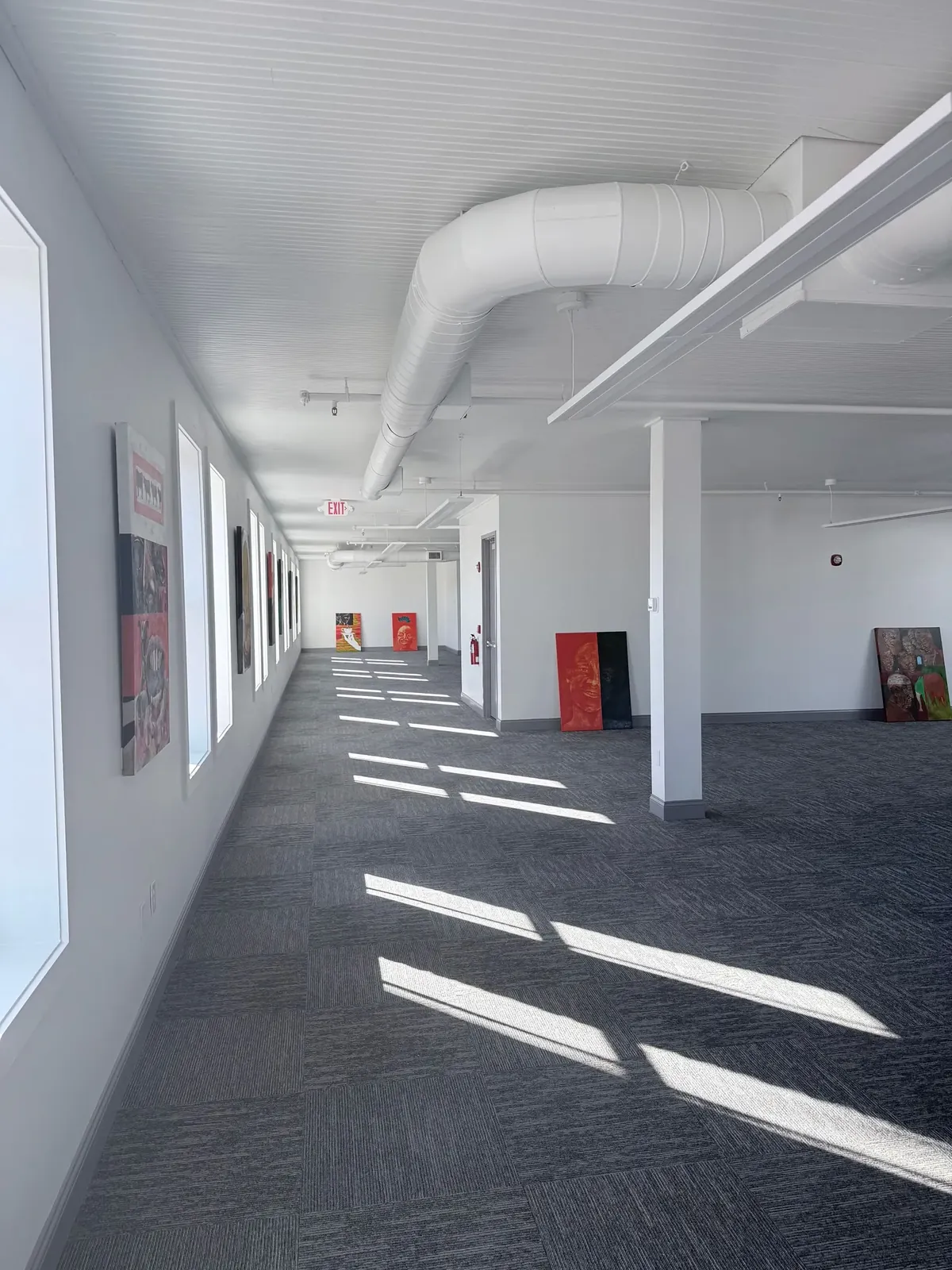 A bright, open-plan office space featuring a gray patterned carpet, white walls, exposed ceiling ducting, and wall art.